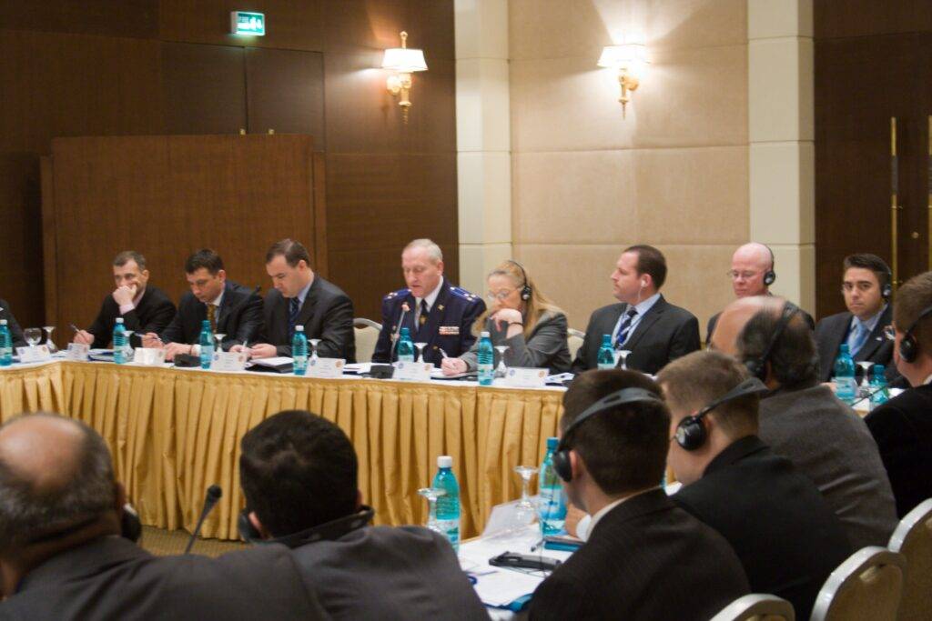 A group of people in formal attire sit around a conference table with microphones and bottled water, engaged in discussion. Some wear headsets, and a man in uniform speaks at the center.