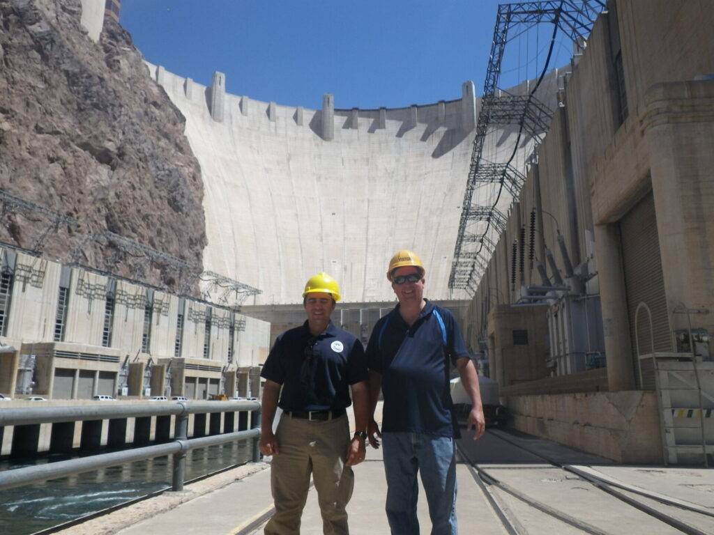 As seen above, two engineers in safety gear stand before the Hoover Dam, illustrating scale and typical site safety protocols.