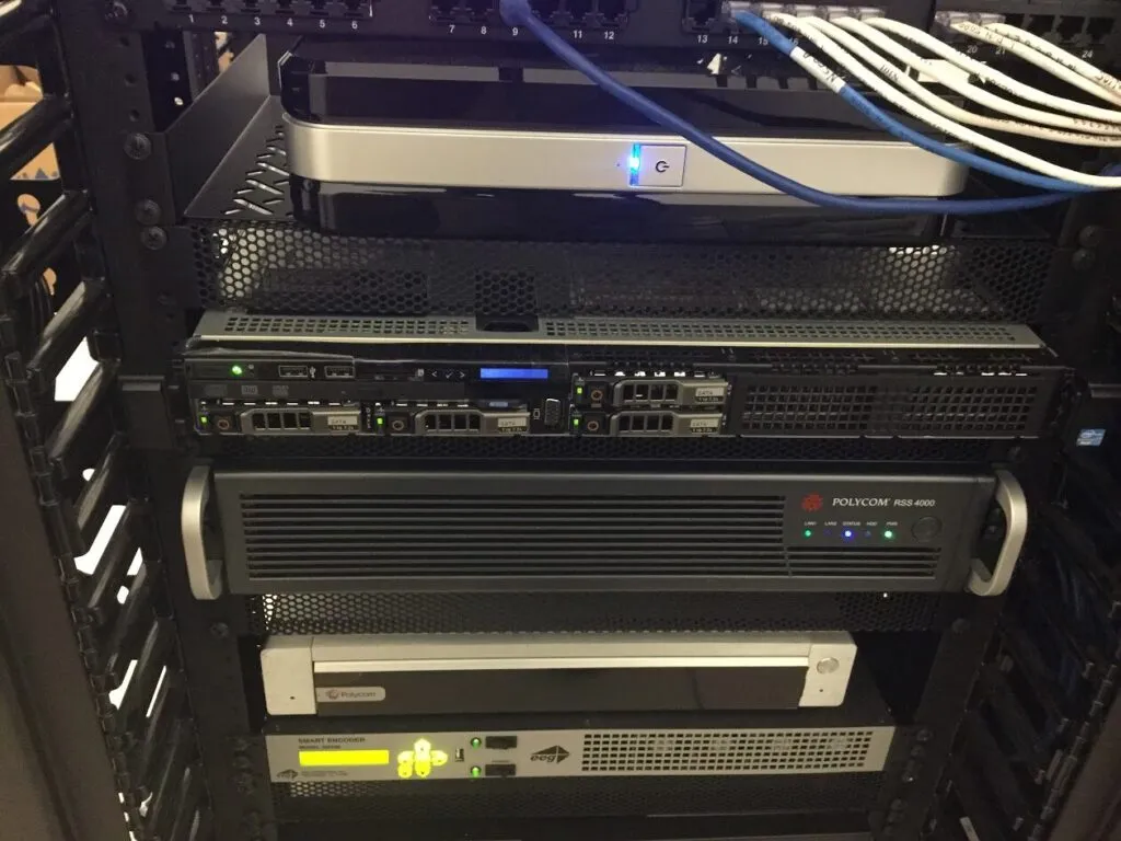 A close-up view of a server rack with various network equipment, including servers and switches, neatly connected with organized network cables. Multiple indicator lights are illuminated on the devices.