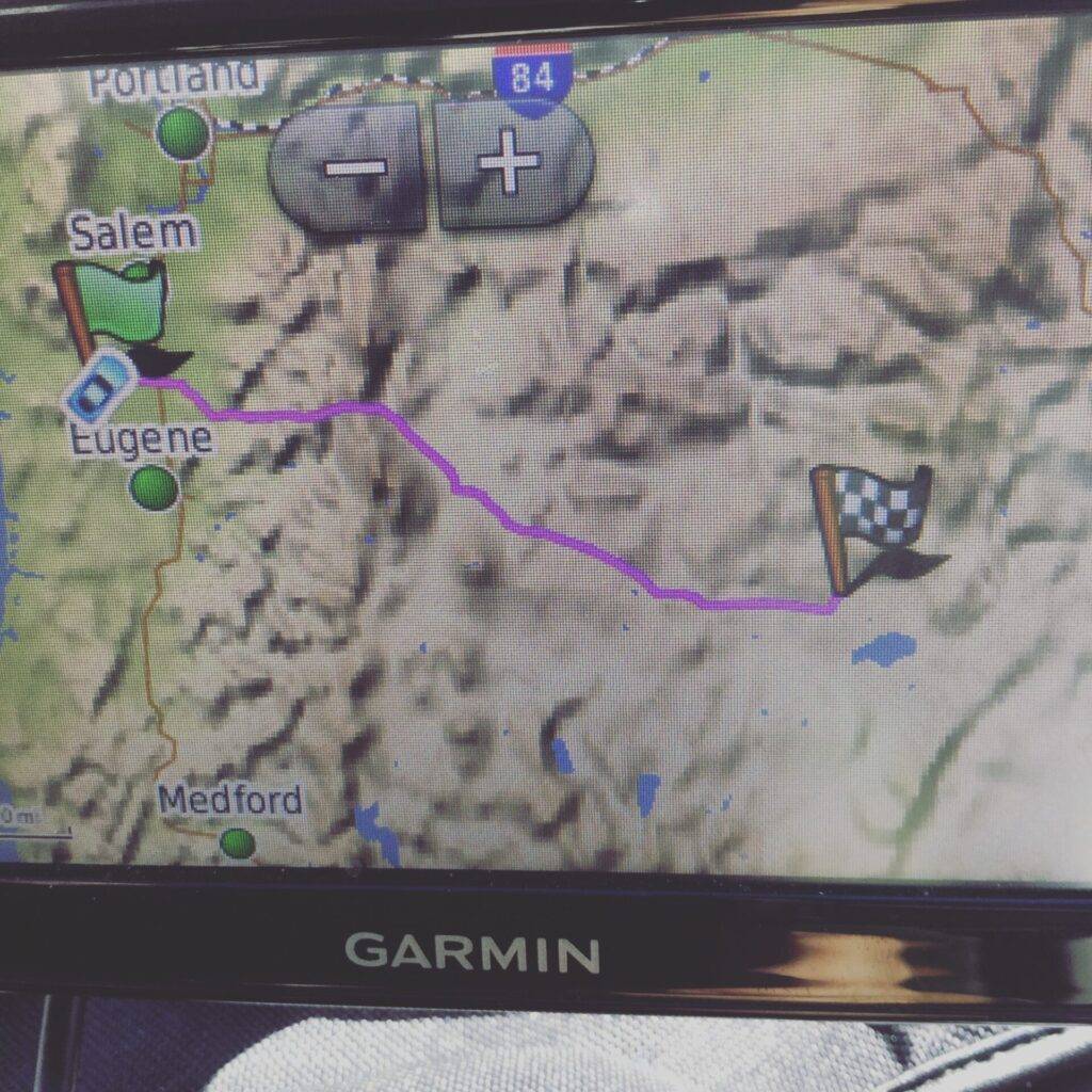 A GPS screen showing a mapped route from Eugene, Oregon, heading east with a purple line to a checkered flag destination. Cities like Portland, Salem, and Medford are also visible on the map.