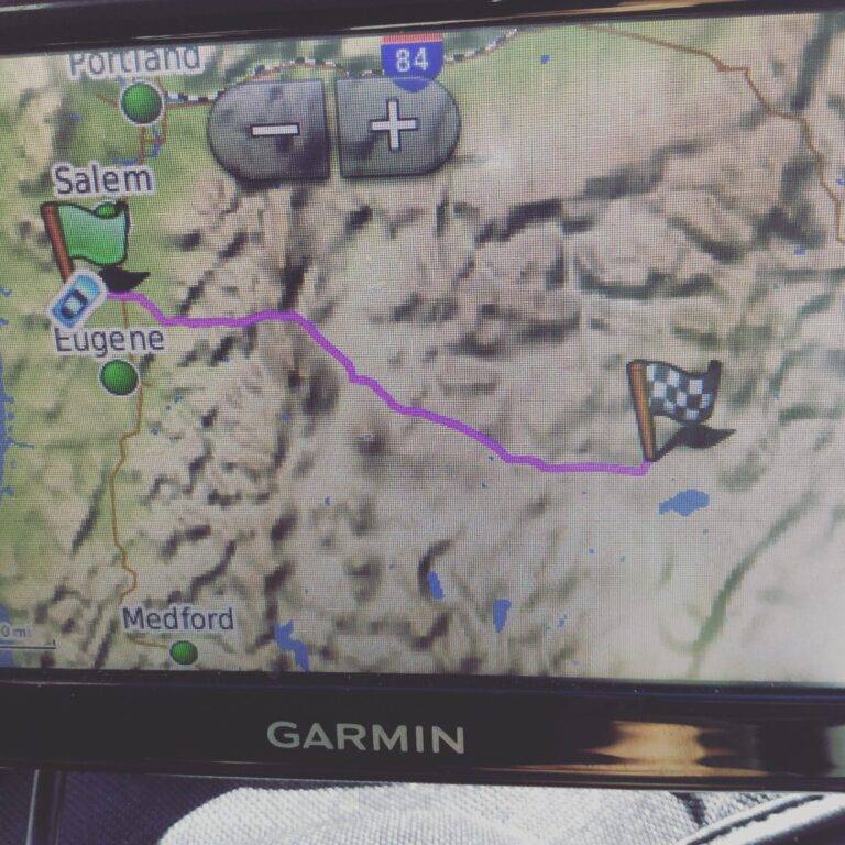 A GPS screen showing a mapped route from Eugene, Oregon, heading east with a purple line to a checkered flag destination. Cities like Portland, Salem, and Medford are also visible on the map.