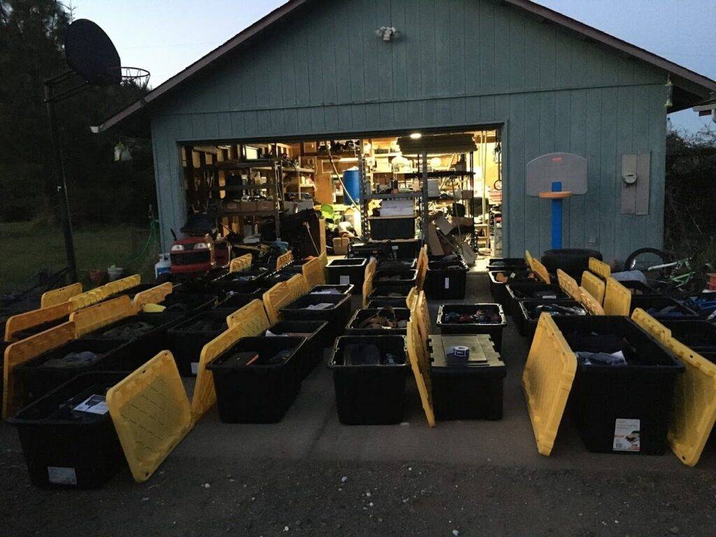Rows of large black storage bins with yellow lids, some open, are arranged in front of a garage with shelves full of tools and equipment, suggesting a large organizing or moving project.