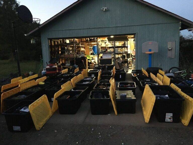 Rows of large black storage bins with yellow lids, some open, are arranged in front of a garage with shelves full of tools and equipment, suggesting a large organizing or moving project.