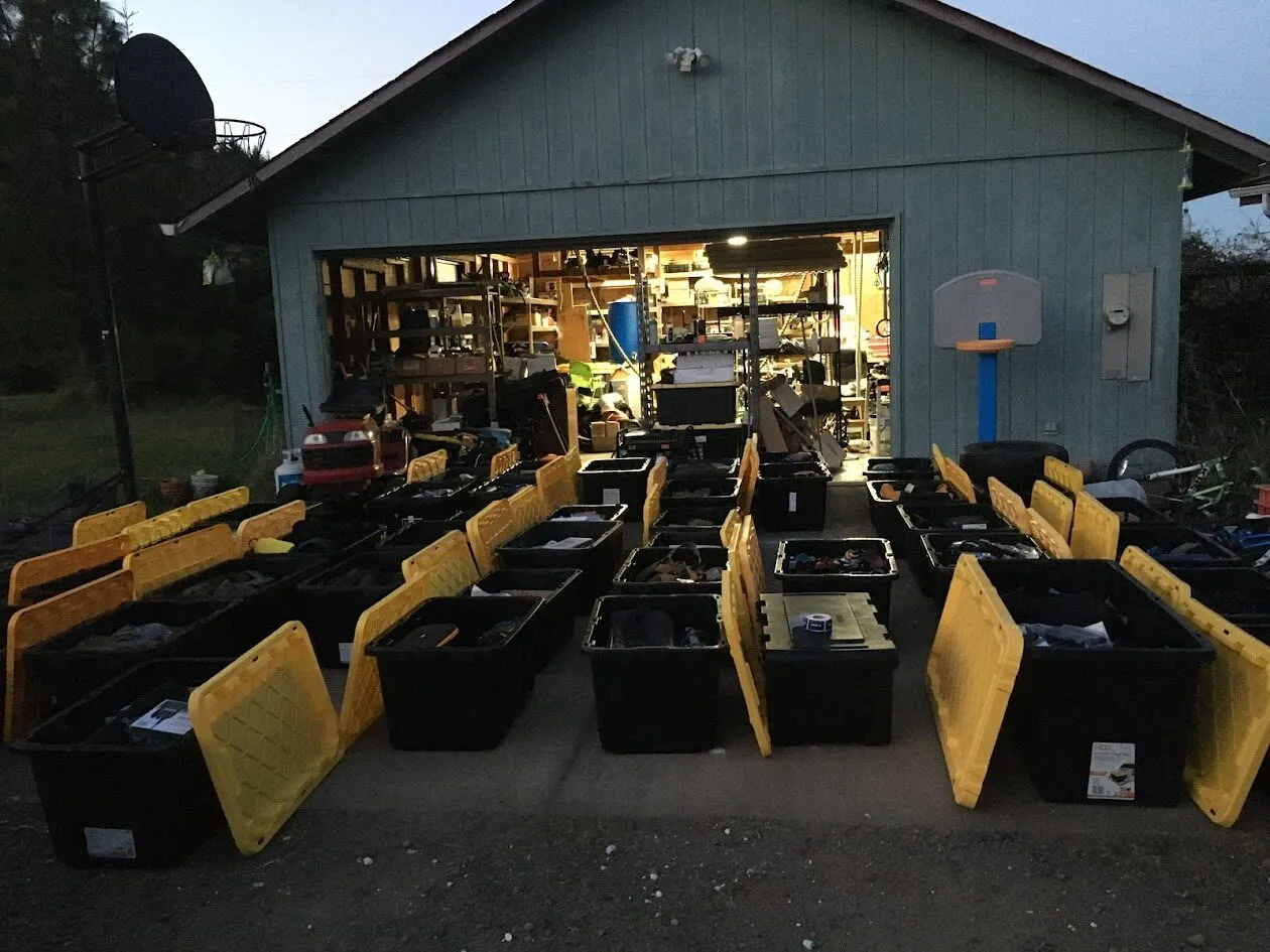 Rows of large black storage bins with yellow lids, some open, are arranged in front of a garage with shelves full of tools and equipment, suggesting a large organizing or moving project.