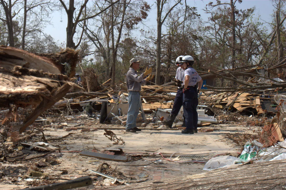 Three people, including two emergency responders in uniform and helmets, talk amid debris and fallen trees in a devastated area after a natural disaster. The surroundings show significant destruction and scattered wreckage.