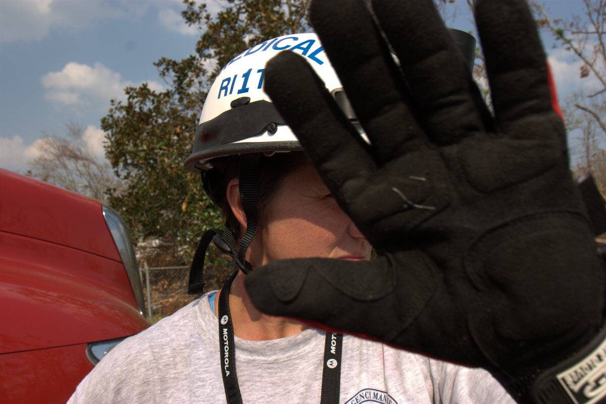 A person wearing a helmet and gloves holds up their hand to block the camera, obscuring most of their face. Trees and a red vehicle are visible in the background.
