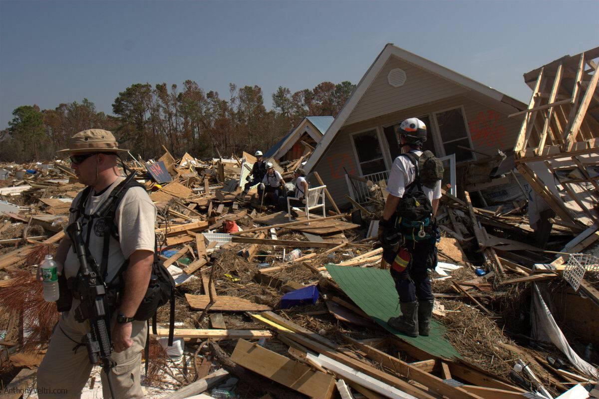 Rescue workers stand among debris and collapsed houses after a natural disaster. They wear protective gear and safety helmets, searching through the wreckage for survivors. Trees and damaged buildings are visible in the background.