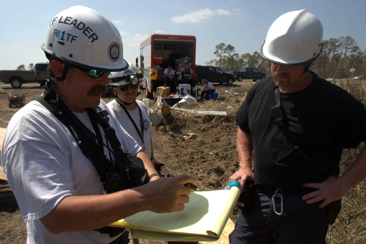Two men in hard hats and safety gear stand outdoors at a work site, one writing on a yellow notepad while the other looks on. Equipment, a trailer, and several people are visible in the background.