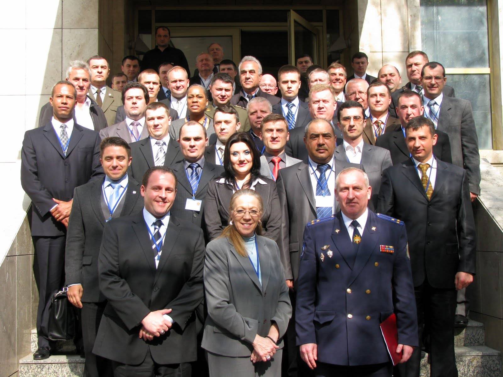 A large group of professionally dressed men and women, some in uniforms, stand together on outdoor steps in front of a building, posing and smiling for a group photo.