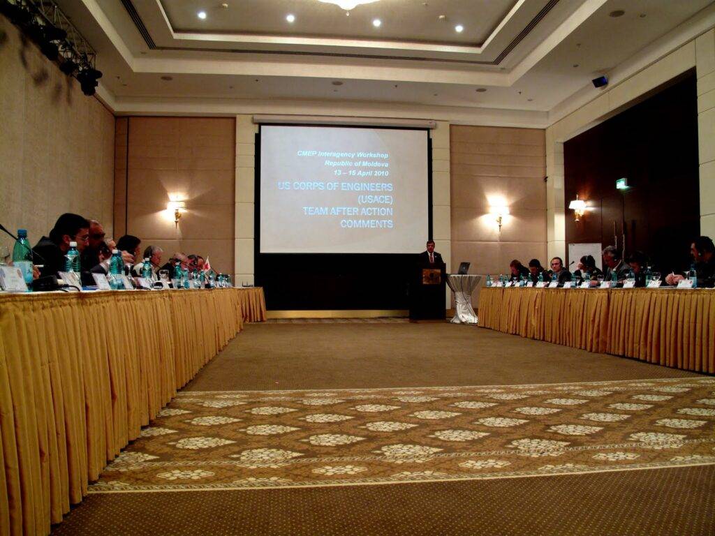 A large conference room with people seated around two long tables facing a screen. A presenter stands at a podium. The screen displays a workshop title and the text "US Corps of Engineers (USACE) Team After Action Comments.