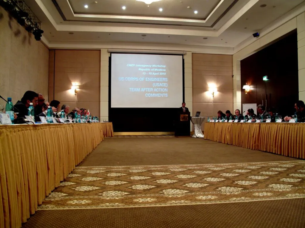 A large conference room with people seated around two long tables facing a screen. A presenter stands at a podium. The screen displays a workshop title and the text "US Corps of Engineers (USACE) Team After Action Comments.