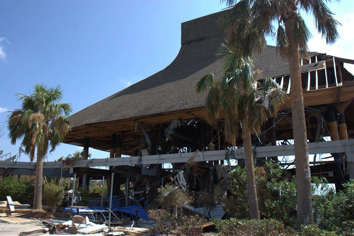 A large, damaged building with exposed beams and debris, surrounded by palm trees and overgrown plants, under a clear blue sky. The structure appears partially collapsed and abandoned.