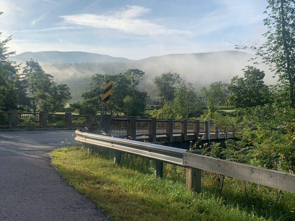 A small bridge with a metal guardrail curves over a stream, surrounded by green trees and grass. Mist rises in the distance, partially obscuring hills under a blue, lightly clouded sky.