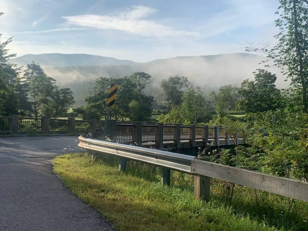A small bridge with a metal guardrail curves over a stream, surrounded by green trees and grass. Mist rises in the distance, partially obscuring hills under a blue, lightly clouded sky.