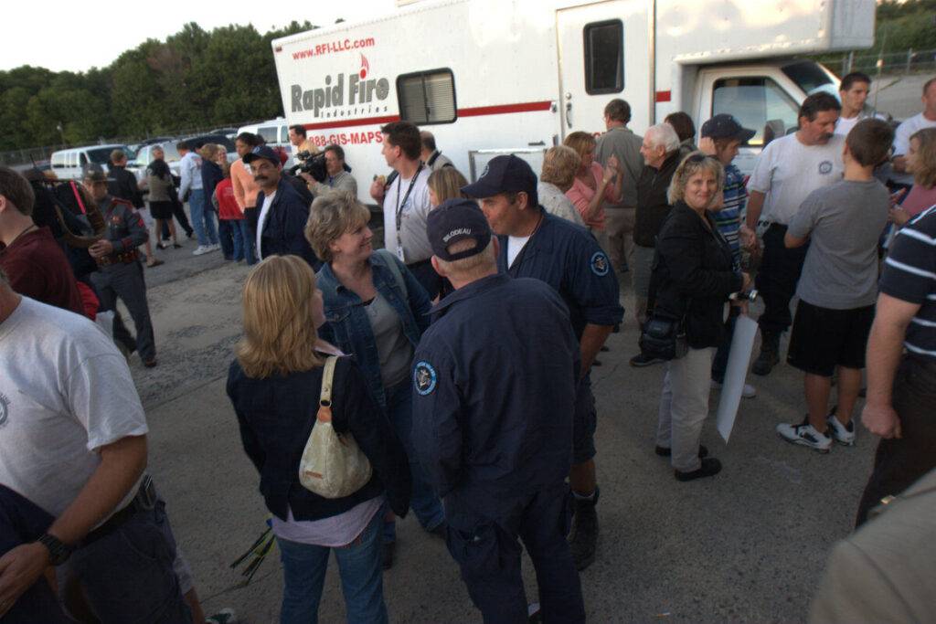 A group of people, including emergency workers in uniform, gather and talk outside near a white "Rapid Fire" truck parked on a paved area. Some people are smiling and engaged in conversation. Trees are visible in the background.