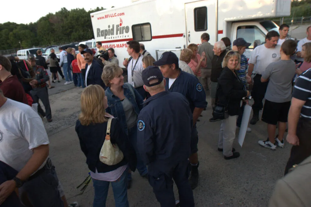 A group of people, including emergency workers in uniform, gather and talk outside near a white "Rapid Fire" truck parked on a paved area. Some people are smiling and engaged in conversation. Trees are visible in the background.