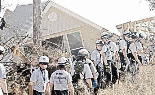 A team of urban search and rescue workers in helmets and uniforms stand in a line near a collapsed house and debris, appearing to coordinate recovery efforts after a disaster.