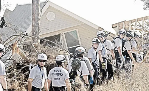 A team of urban search and rescue workers in helmets and uniforms stand in a line near a collapsed house and debris, appearing to coordinate recovery efforts after a disaster.