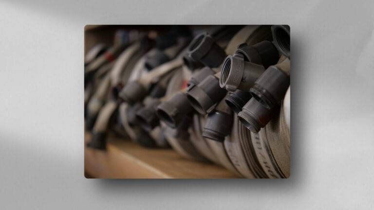 A close-up of several coiled fire hoses with metal couplings, stacked in a row on a shelf. The image is focused on the connectors, with the hoses extending into a blurred background.