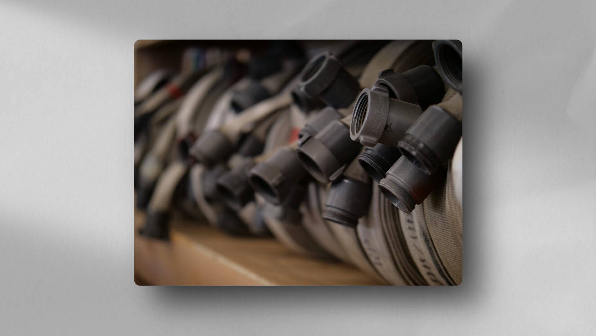 A close-up of several coiled fire hoses with metal couplings, stacked in a row on a shelf. The image is focused on the connectors, with the hoses extending into a blurred background.