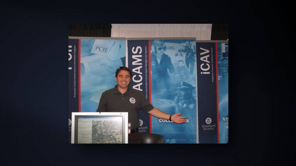 A person stands smiling with arms outstretched at a booth with Homeland Security banners and a computer monitor in the foreground. The background features various informational posters and security-related graphics.