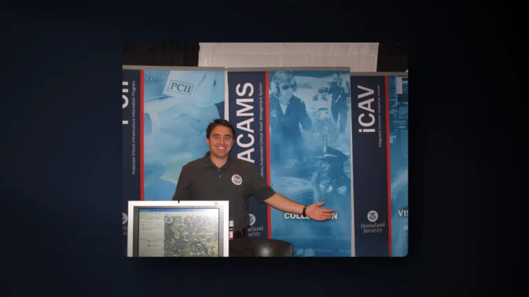 A person stands smiling with arms outstretched at a booth with Homeland Security banners and a computer monitor in the foreground. The background features various informational posters and security-related graphics.
