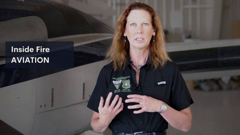 Forest Service lead plane pilot Mary Verry speaking in a hangar with a King Air aircraft behind her.