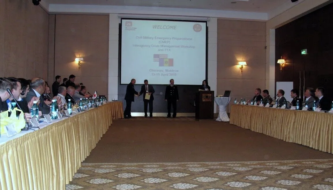 A conference room with people seated along two long tables facing a screen. Four people stand at the front near a podium. The screen displays a welcome message for an emergency preparedness workshop in Chisinau, Moldova.