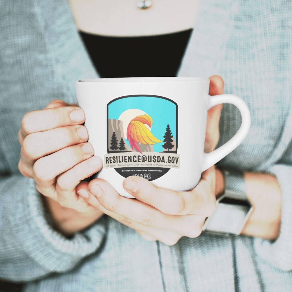 A person in a gray sweater holds a white mug with a colorful logo showing a sunrise, trees, and mountains, and the text “RESILIENCE@USDA.GOV” on the front.