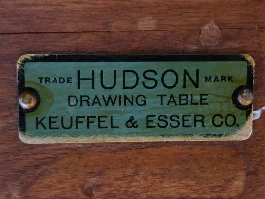 This image presents a worn green metal label on wood, identifying a Keuffel & Esser Co. drawing table by its trade mark.