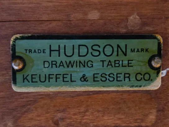 This image presents a worn green metal label on wood, identifying a Keuffel & Esser Co. drawing table by its trade mark.