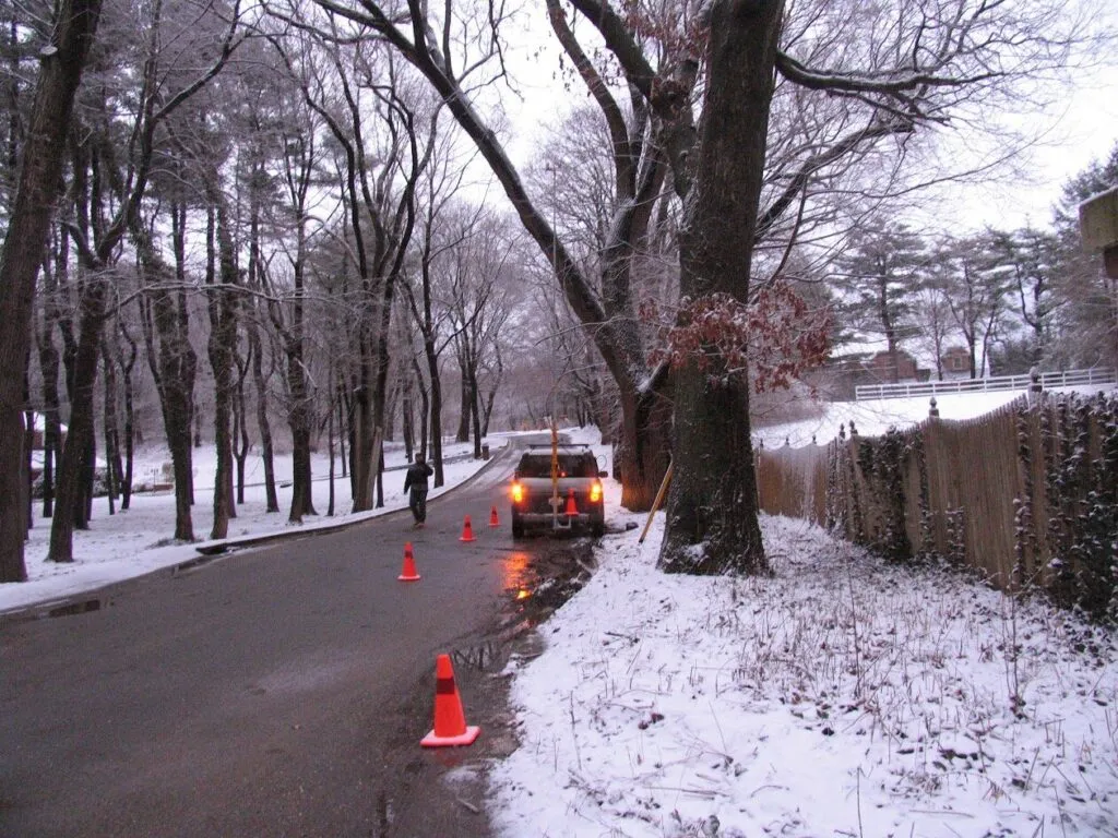 The image illustrates a snow-covered road with safety cones and hazard lights, suggesting caution is needed in winter conditions.