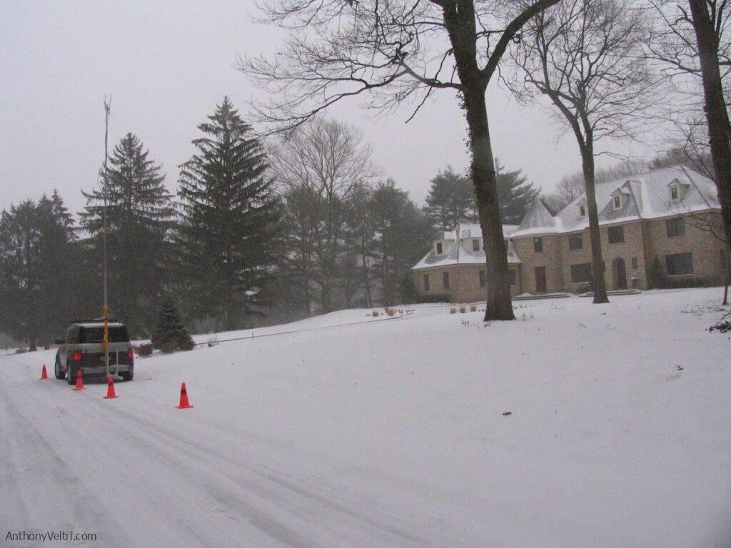 The image depicts a snow-covered street, where a parked vehicle and traffic cones may indicate ongoing utility or survey work.