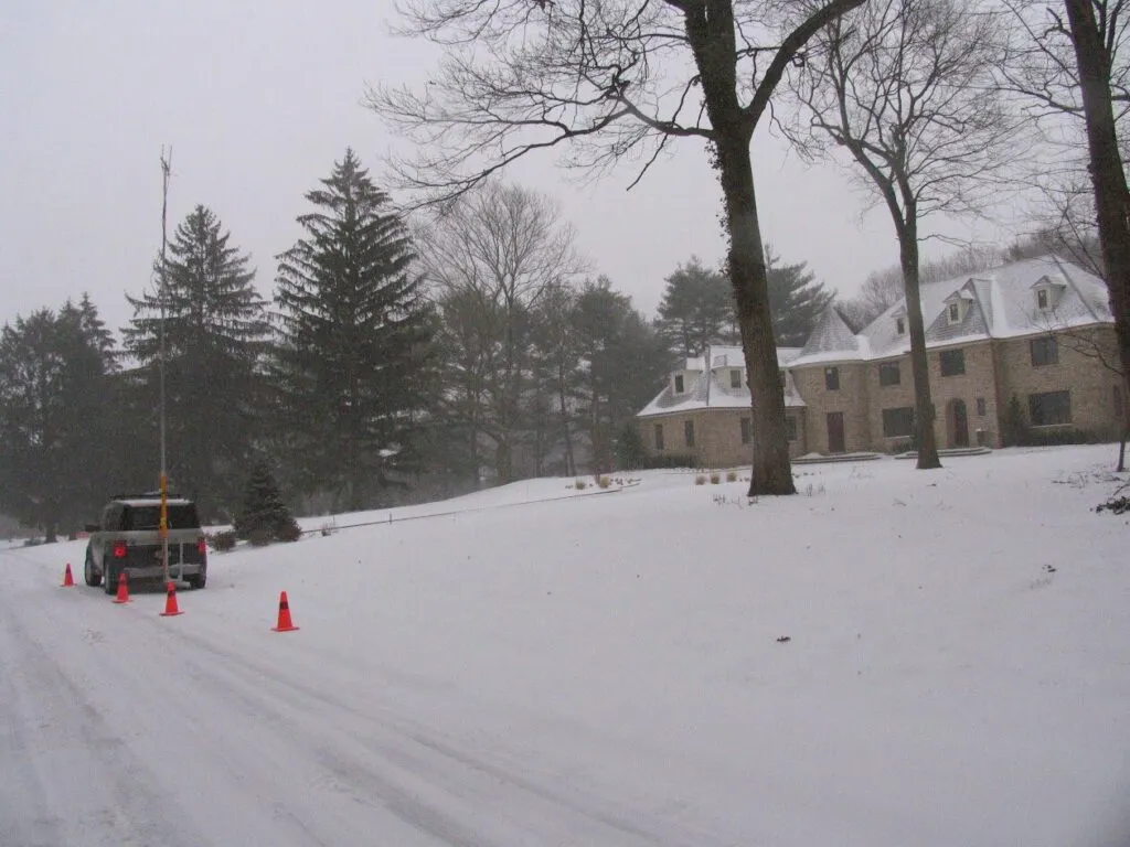 The image depicts a snow-covered street, where a parked vehicle and traffic cones may indicate ongoing utility or survey work.