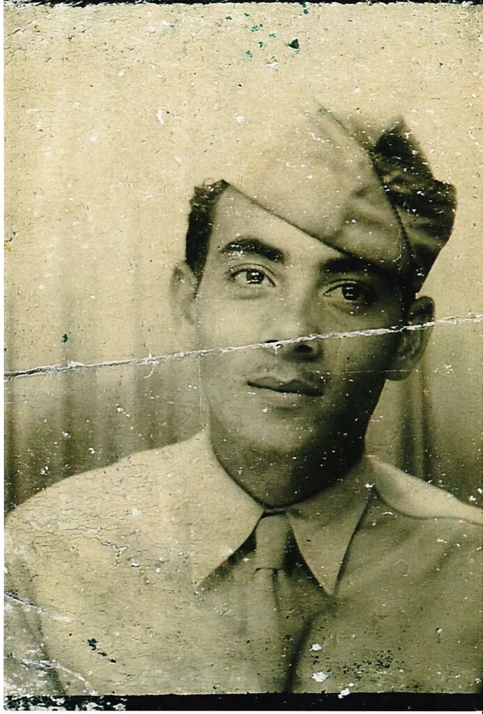 A vintage, sepia-toned portrait of a man in a military uniform and cap, gazing slightly upward. The photo is aged, with visible creases and small marks across its surface.