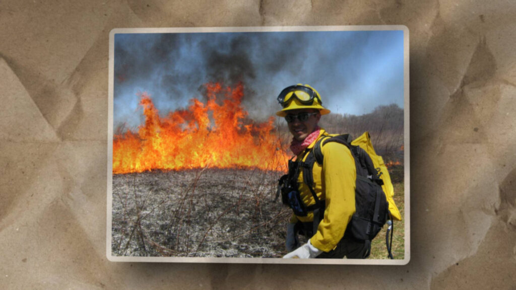 A firefighter in yellow protective gear stands in front of a blazing wildfire, with smoke rising into the sky and dry grass burning intensely behind him.