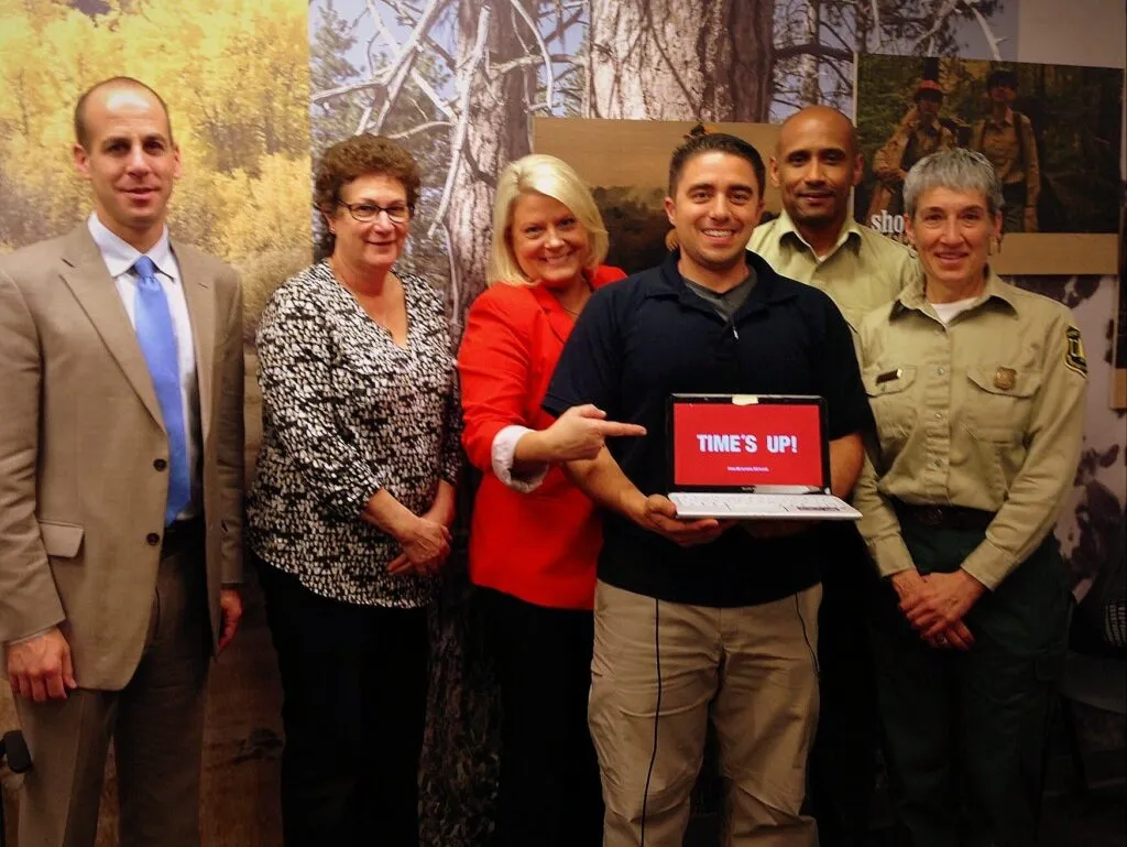 Six adults stand indoors, smiling at the camera. The man in the center holds a laptop displaying "TIME'S UP!" in red and white. They are dressed in business or uniform attire with a woodsy, nature-themed background.