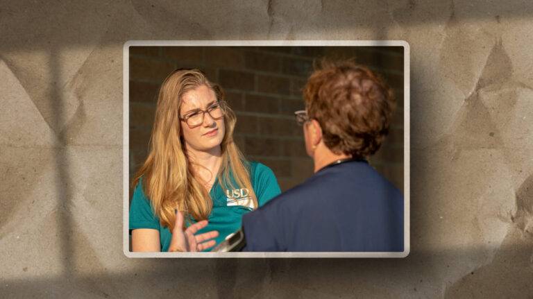A woman with long blonde hair and glasses listens intently to a man while talking outdoors. The woman wears a teal shirt, and the background features a brick wall with a paper-like border around the image.