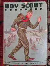 Cover of a vintage Boy Scout Handbook shows a smiling scout in uniform carrying a backpack, saluting as he walks. Illustrated scouts, tents, a campfire, and trees are in the background. Title reads “Boy Scout Handbook. Boy Scouts of America.”.