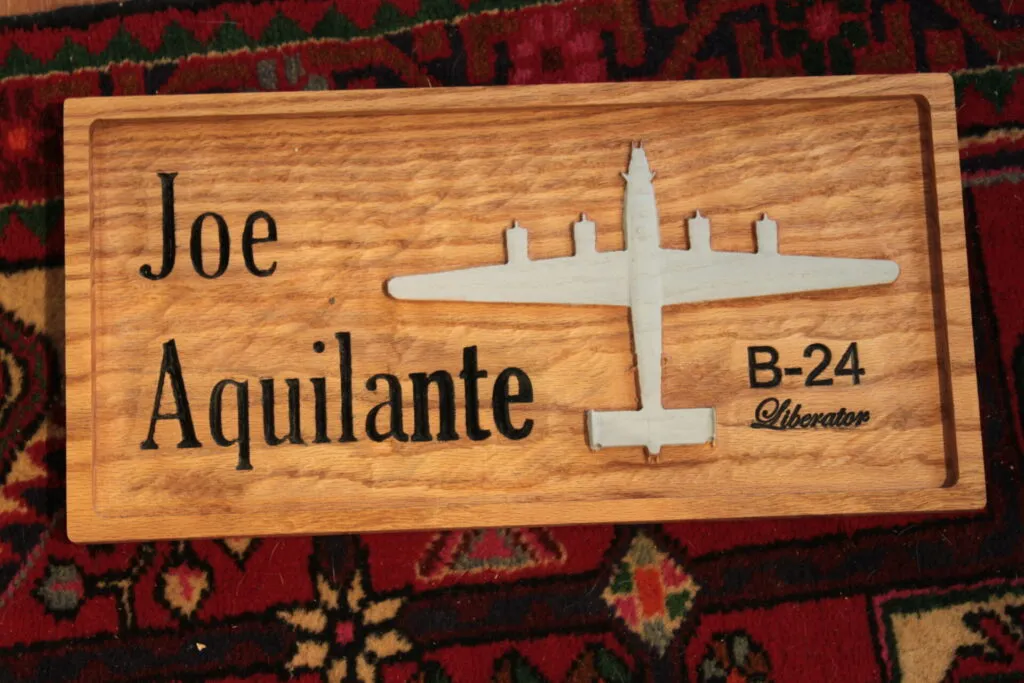 A wooden plaque with “Joe Aquilante” on the left, and a silhouette of a B-24 Liberator bomber on the right above the text “B-24 Liberator.” The background is a patterned red and green carpet.