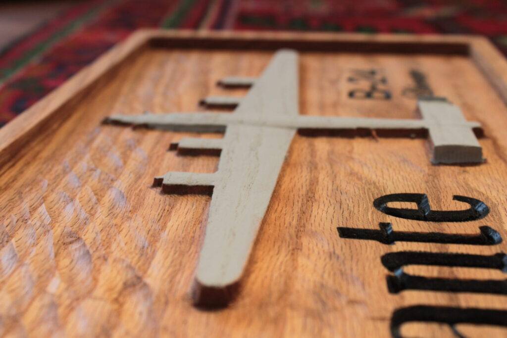 A wooden plaque features a carved silhouette of a four-engine airplane viewed from above. The plane is painted beige, and black engraved text is partially visible on the right. The background is a reddish patterned surface.