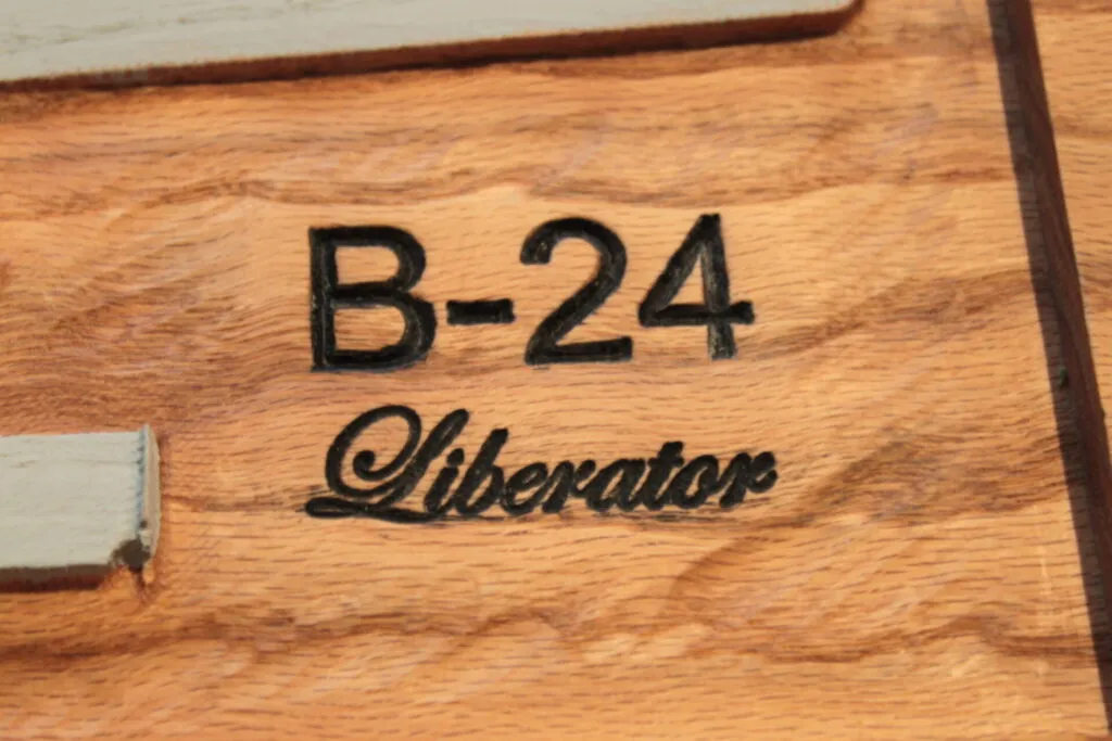 Close-up of wood with "B-24 Liberator" carved and painted in black. The grain of the wood is visible, and part of a gray painted section appears at the top and left edges. The style is rustic and hand-crafted.