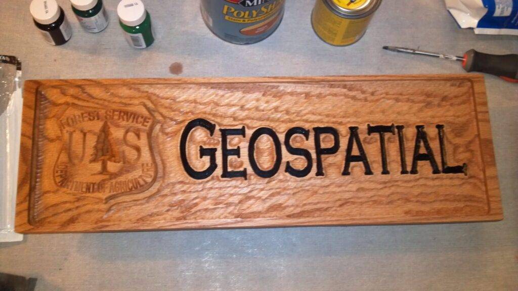 A wooden plaque reads "GEOSPATIAL" in large, bold black letters. On the left, there is a carved USDA Forest Service shield. Paint cans, tools, and a small green bottle sit on the gray surface around the plaque.