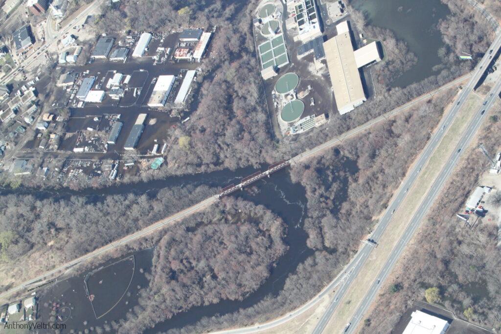 Aerial view of a tree-covered river area crossed by a bridge, with a circular land formation in the center. Industrial and utility buildings are visible to the upper left and right; a multi-lane road runs parallel to the river at the bottom right.