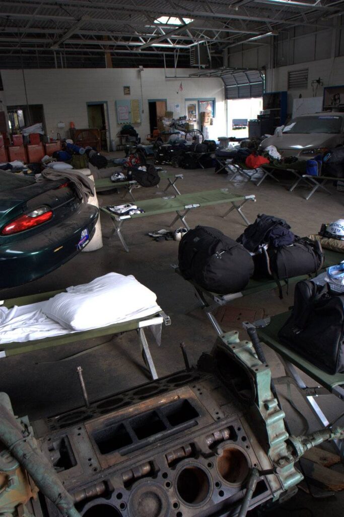 Military-style cots with bedding and bags are arranged in an automotive garage. Cars, tools, and engine parts are visible. The setting has high ceilings, fluorescent lights, and posters on the walls, blending a workshop with a makeshift sleeping area.
