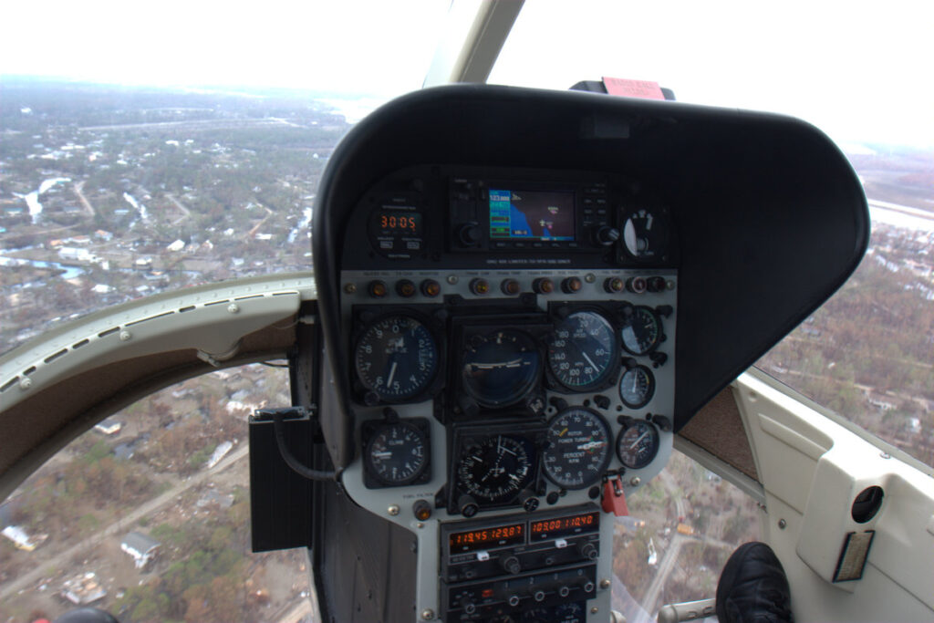 A cockpit view from a helicopter flying over a landscape, showing various flight instruments, gauges, and controls on the control panel, with the pilotโs gloved hand visible on the right and a blurry, wooded area outside the windows.