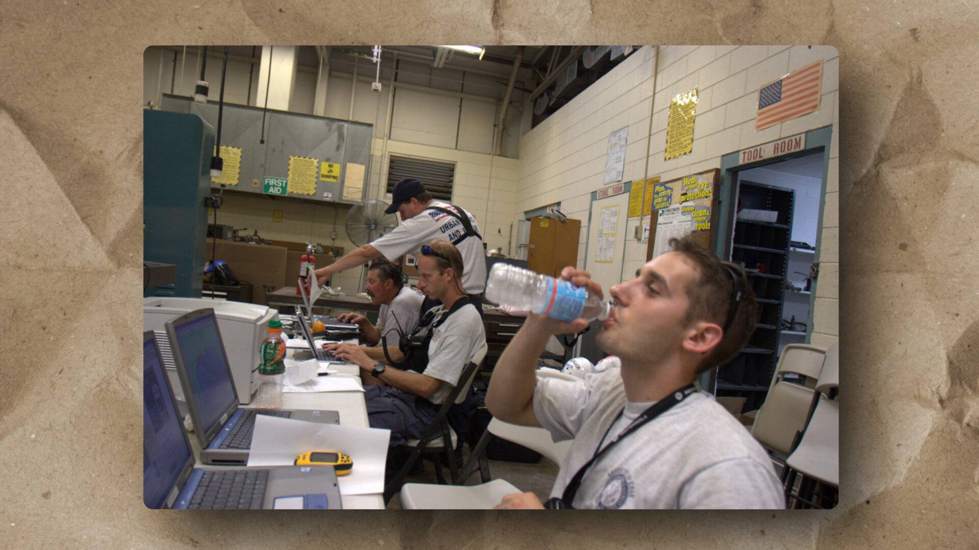 Four people sit at a table with laptops, papers, and drinks in a room with bulletin boards and an American flag. One person stands, reaching for something on the table, while another drinks from a water bottle in the foreground. The setting appears to be an office or control room.