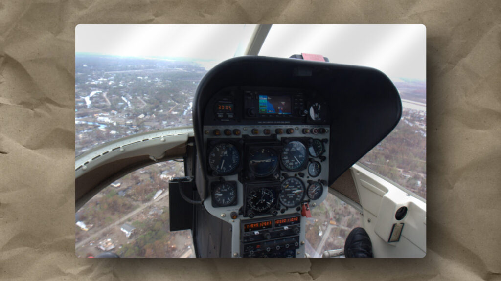 Aerial view from inside a helicopter cockpit, showing detailed instrument panels with multiple dials and screens. Through the windshield, a landscape with roads, buildings, and trees is visible. A pilot's foot rests on the control pedal in the lower right corner.