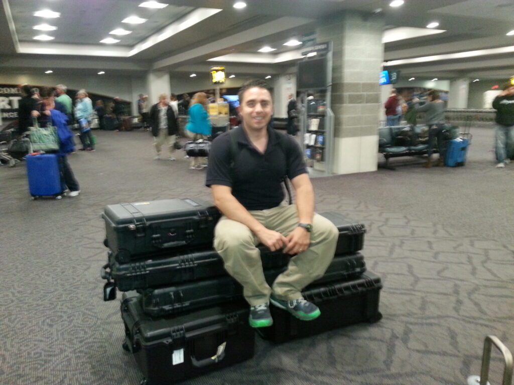 A man in a black polo and khaki pants sits smiling on stacked black hard cases in an airport terminal. People with luggage are seen in the background near gates. The setting is busy with bright overhead lighting and gray carpeting.