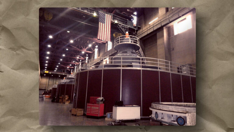 Interior of a large industrial facility with several massive, cylindrical machines lined up. An American flag hangs above them. The area is lit by rows of ceiling lights and scattered with equipment, boxes, and railings, giving a sense of scale and function.